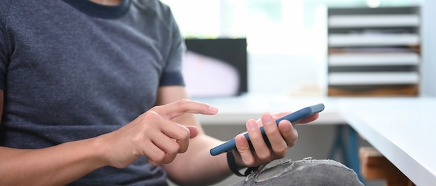 Close-up van jonge man hand typen op het scherm van slimme telefoon zittend op zijn werkplek.