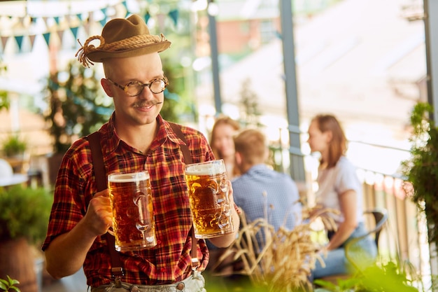 Foto close-up van jonge man in geruit overhemd en fedora hoed bier drinken wazig mensen op de achtergrond