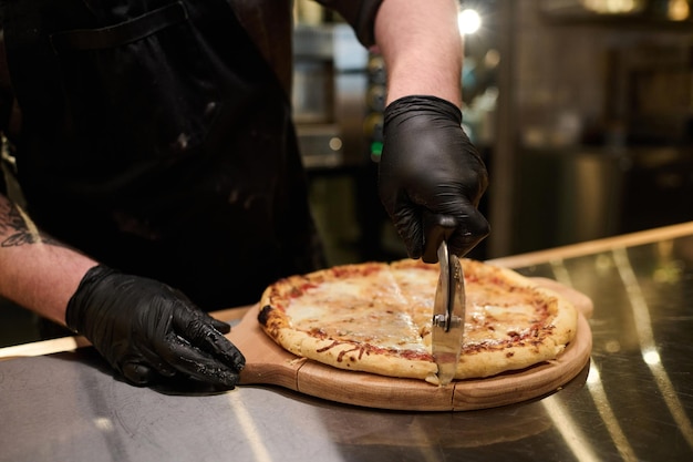 Close-up van jonge mannelijke kok in handschoenen die smakelijke pizza snijden