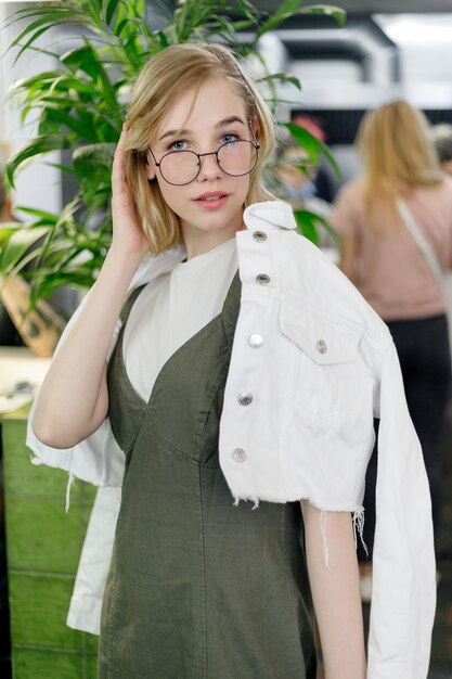 Foto close-up van jonge vrolijke vrouw in jurk die gelukkig in de camera kijkt met grote groene bladeren op de achtergrond in een gezellig groen café. meisje met bril