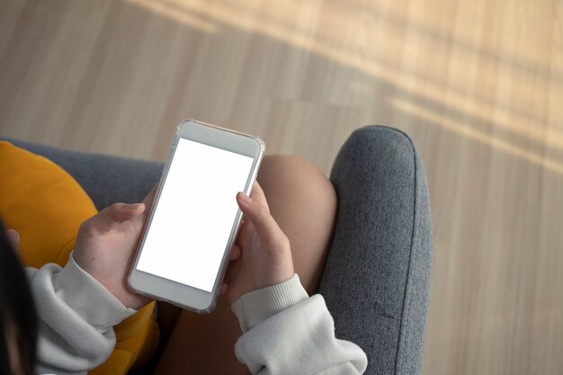 Close-up van jonge vrouw die mock-up smartphone met leeg scherm vasthoudt terwijl ze thuis op de bank zit.