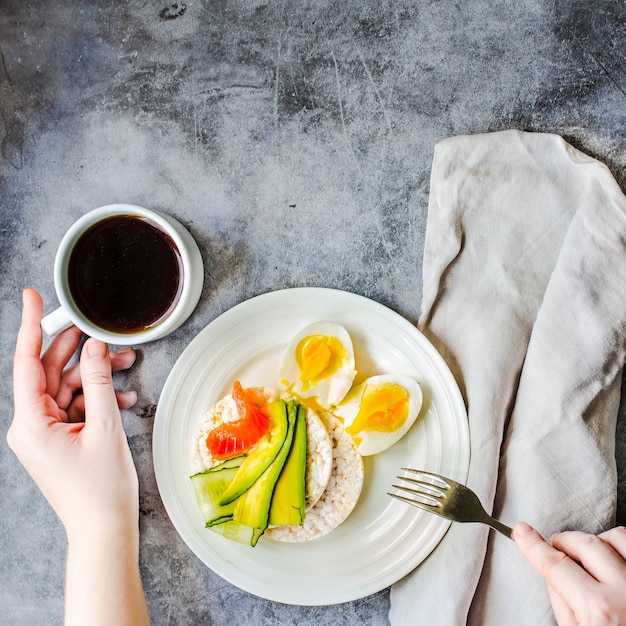 Foto close-up van jonge vrouw met een lunchpauze. rijstkrokante cakejes met avocado en vers gezouten zalm. mannelijke handen met voedsel dat rijk is aan vet en eiwit en kopje koffie. bovenaanzicht. ruimte voor tekst.