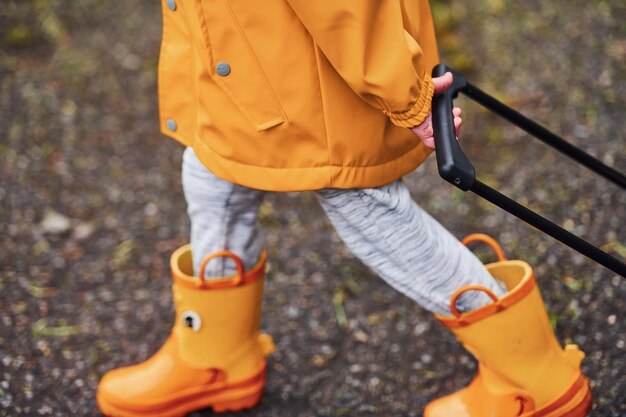 Foto close-up van kind in gele waterdichte mantel en laarzen die buiten lopen met koffer