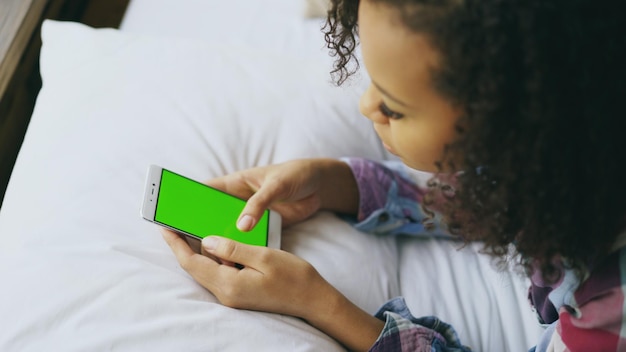 Close-up van krullend gemengd ras vrouw liggend in bed thuis met behulp van smartphone met groen scherm
