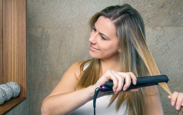 Foto close-up van lachende jonge vrouw die haar haar rechttrekt met een stijltang in de badkamer. gezondheid en schoonheidsconcept.