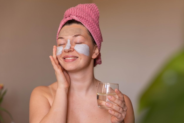 Close-up van lachende vrouw met cosmetisch masker op gezicht met glas water