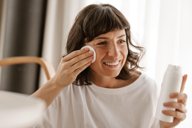 Close-up van lachende vrouw met lotion voor gezicht kijk naar spiegel