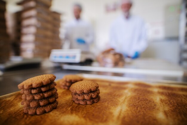 Close up van lade vol met vers gebakken koekjes in voedselfabriek. Wazig beeld van twee mannelijke werknemers in steriele kleren op de achtergrond.