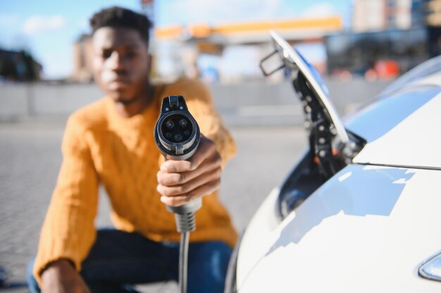 Close up van mannenhand met elektrische oplaadstekker voor luxe rode auto. Afrikaanse zakenman laadt zijn auto op station. Milieuvriendelijk vervoer.