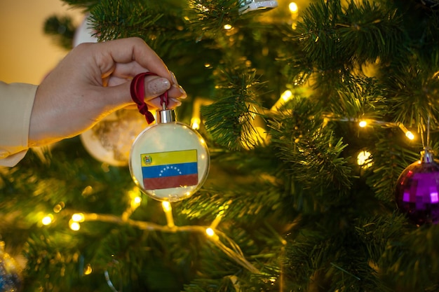 Close up van meisjes hand met een kerstbal voor een dennenboom met de vlag van Venezuela Nieuwjaar in Venezuela kerstvakantie wenskaart met kopie ruimte Huis versieren voor viering