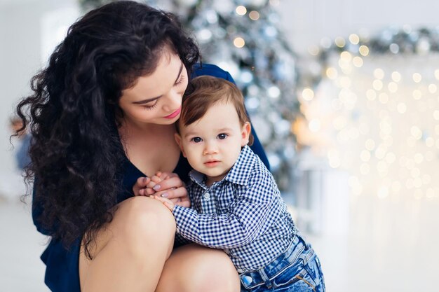 close-up van moeder kijkt naar kleine zoon en houdt zijn handen vervaagd kerstboom en kransen op de achtergrond