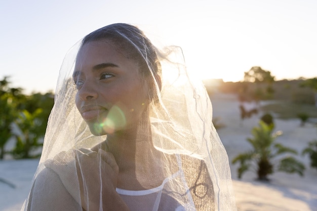 Foto close-up van mooie biracial jonge bruid die sluier draagt en wegkijkt naar het strand tegen heldere hemel