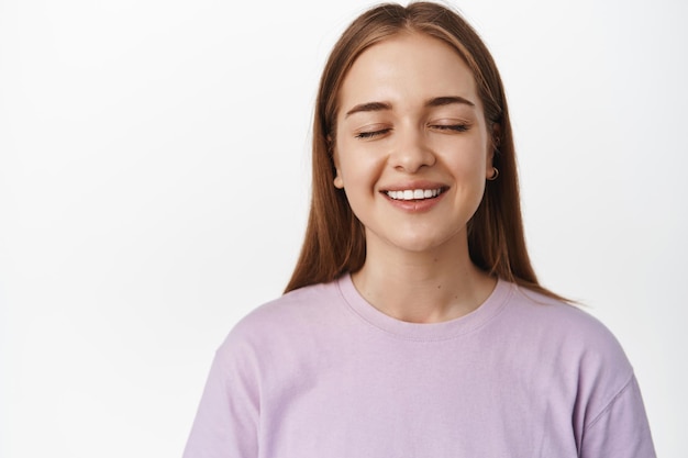 Close-up van mooie jonge vrouw zonder make-up, natuurlijk gezicht, gesloten ogen dromerig en glimlachen, iets in beeld brengen, wensen doen of dromen, staande op een witte achtergrond.