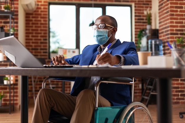 Close up van ondernemer met een handicap die met laptop werkt aan bedrijfsstrategie. Man met gezichtsmasker zittend in een rolstoel om te werken aan projectplanning op het kantoor van het opstartende bedrijf.