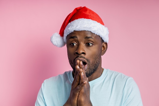 Close-up van ongeschoren verraste afro-amerikaanse man in kerstmuts, kijkt naar de kant met geschokte uitdrukking, ziet iets ongelooflijks, opent zijn mond van verbazing op roze achtergrond. kerstconcept.