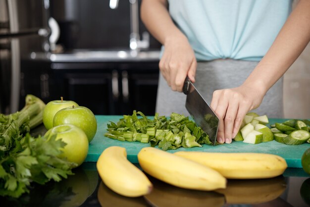 Close-up van onherkenbare vrouw die aan de balie staat met groenten en fruit en selderij snijdt op plastic bord