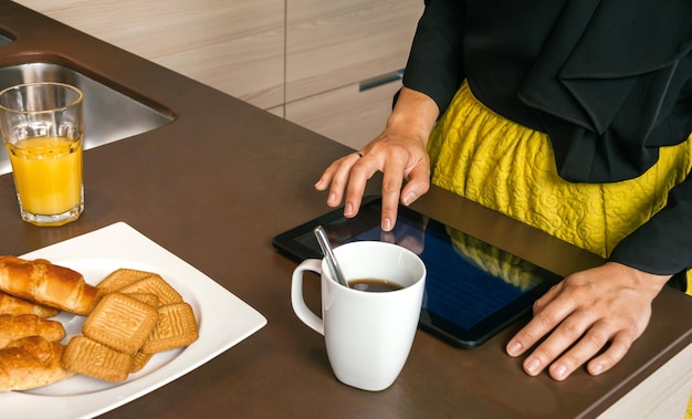 Close-up van onherkenbare vrouw klaar om uit te gaan met behulp van elektronische tablet terwijl ze snel ontbijt in de keuken