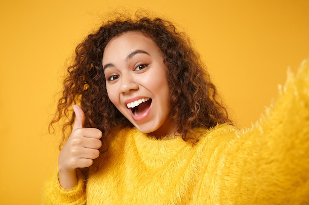 Close up van opgewonden jonge Afro-Amerikaanse meisje in trui poseren geïsoleerd op gele muur achtergrond. Mensen levensstijl concept. Bespotten kopie ruimte. Selfie geschoten op mobiele telefoon doen, duim opdagen.