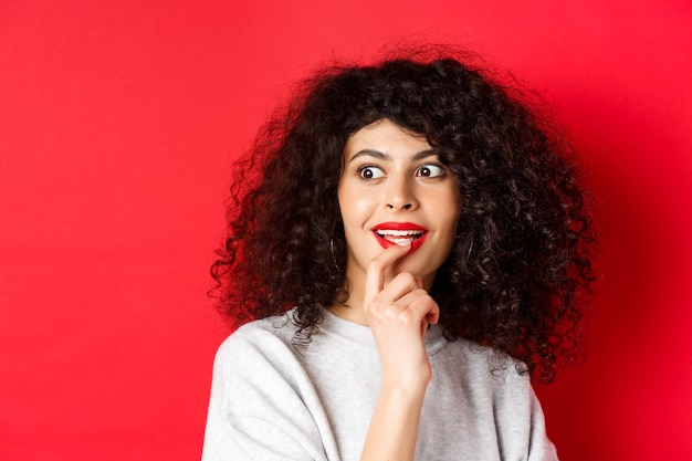 Close-up van opgewonden krullende vrouw die lippen aanraakt en met belangstelling opzij kijkt, coole promo bekijkt, staande tegen een rode muur.