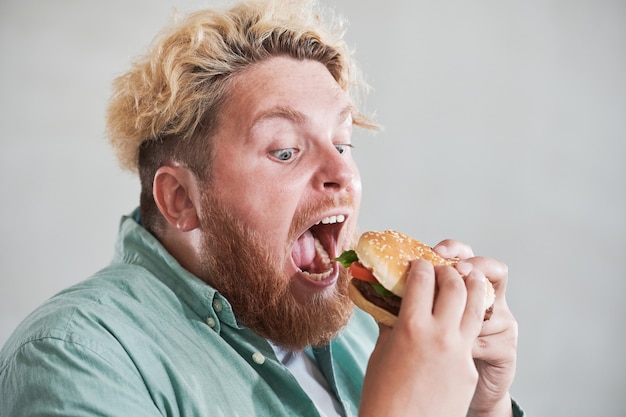 Close-up van overgewicht man die zijn mond opent en grote hamburger eet met plezier geïsoleerd op witte ba...