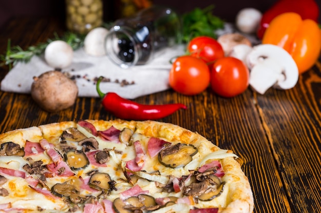 Close up van pizza met ham, ui en augurken op houten tafel, tomaten, champignons en andere groenten op de achtergrond