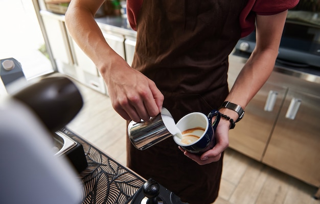 Close-up van professionele barista die gestoomde melk in koffiekop giet en mooie latte achter de toog maakt