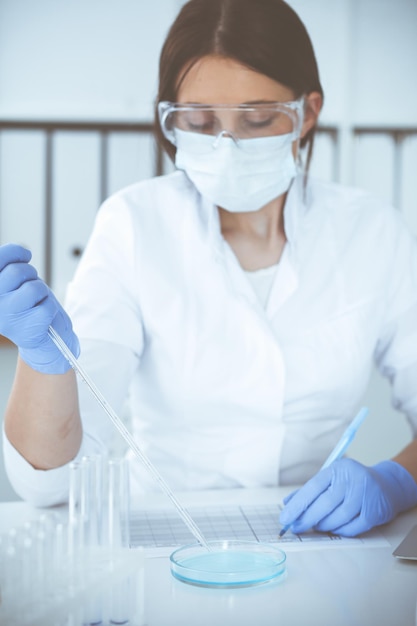 Close-up van professionele vrouwelijke wetenschapper in beschermende brillen die experimenteren met reagentia of bloedonderzoek in het laboratorium. Geneeskunde, biotechnologie en onderzoeksconcept.