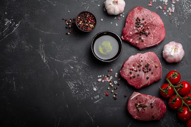 Close-up van rauw vers gemarmerd vlees steak op donkere rustieke betonnen achtergrond met kruiden, olijfolie, knoflook, tomaten klaar om te worden gekookt. Koken vlees filet concept met ruimte voor tekst, bovenaanzicht