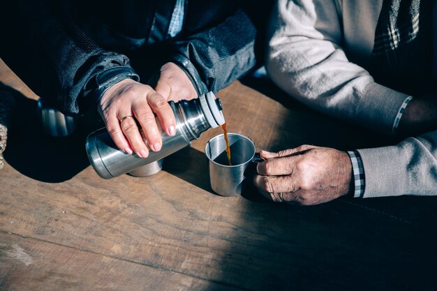 Close up van senior paar handen gieten koffie in een metalen beker uit thermos over een houten tafel