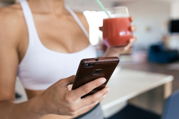 Foto close-up van sportieve jonge vrouw die haar mobiele telefoon gebruikt terwijl ze thuis aardbeiensmoothie drinkt.