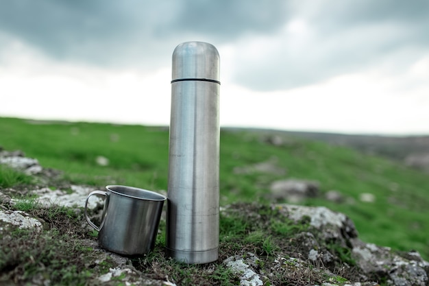 Close-up van stalen roestvrijstalen thermoskan en beker op steen in groen veld.