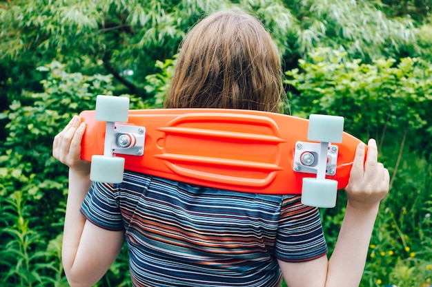 Foto close-up van stijlvol meisje met oranje pennyboard op haar schouders met groene parkachtergrond