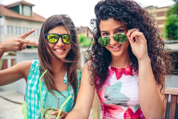 Close-up van twee gelukkige jonge vrouwen met een zonnebril die lacht en naar de camera kijkt terwijl ze buiten groene groentesmoothies houden