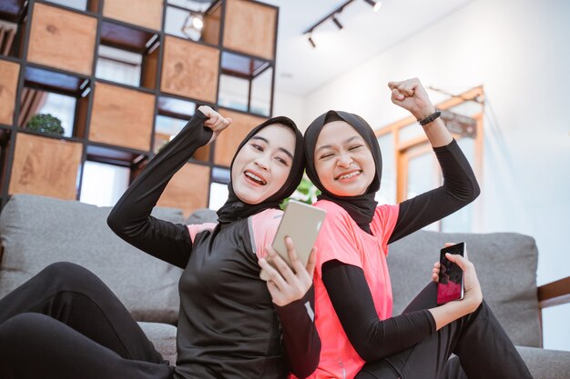 Close-up van twee meisjes die hijab-sportkleding dragen met behulp van een mobiele telefoon terwijl ze op de vloer in het huis zitten