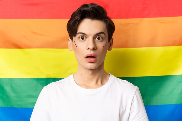 Close-up van verbaasde queer persoon met glitter op gezicht, verbaasd kijken naar camera, staande over lgbtq regenboogvlag.