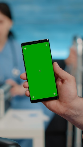 Close-up van verpleegster die verticaal telefoon met groen scherm toont aan senior patiënt die in verpleeghuis woont. Medisch specialist met smartphone met chroma key en geïsoleerde mockup-sjabloon