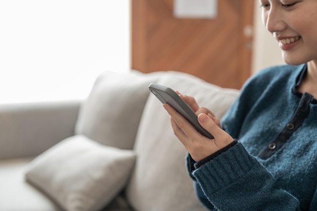 Close-up van vrolijke jonge Aziatische vrouw die mobiele telefoon gebruikt terwijl ze thuis op een bank zit