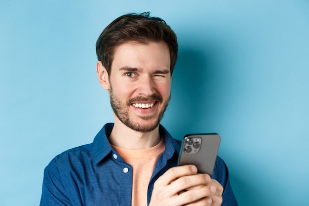 Close up van vrolijke man knipogen en glimlachen, met behulp van mobiele telefoon op blauwe achtergrond.