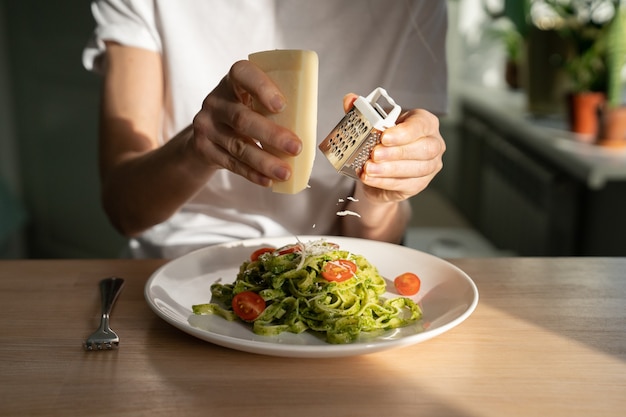 Close up van vrouw handen raspen Parmezaanse kaas in pasta met pesto saus, verse kerstomaatjes, woonkamer op achtergrond.