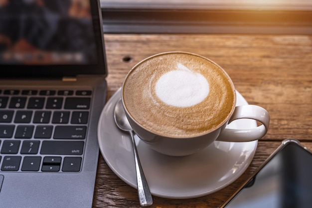 Close-up van warme koffie latte met latte art melkschuim in cup mok en laptopcomputer en smartphone op houten bureau bureau in coffeeshop bij het cafeduring zakelijke werkconcept