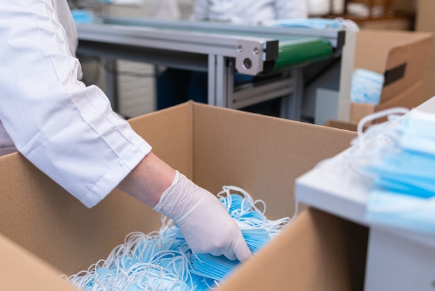 Foto close-up van werknemershanden die handschoenen dragen en medische gezichtsmaskers inpakken in een fabriek.