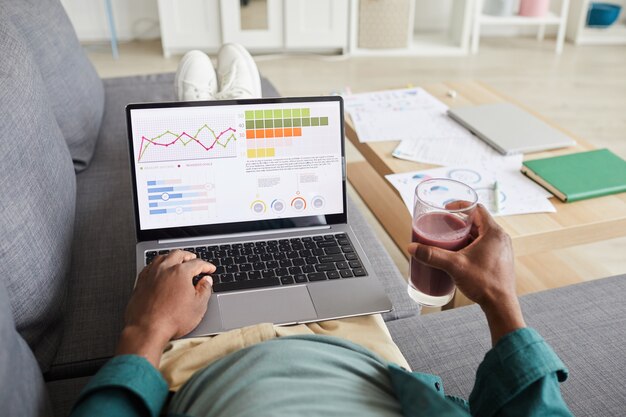 Close-up van zakenman liggend op de bank met verse cocktail en zakelijke grafieken op de monitor van laptop thuis te onderzoeken