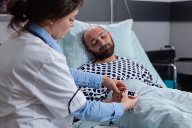 Close up van zieke man patiënt liggend in bed met medische oximeter op vinger. Vrouw arts die hart pluse controleert die ziektecardiogram controleert die in de ziekenhuisafdeling werkt en ziektediagnose uitlegt