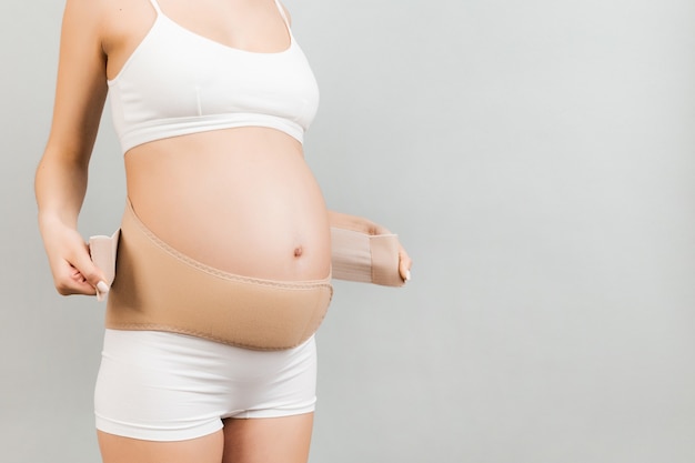 Close up van zwangere vrouw in ondergoed dressing elastische moederschap band op grijze achtergrond met kopie ruimte. Orthopedisch buiksteunriemconcept.