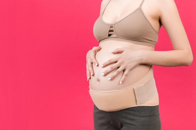 Close up van zwangerschapsverband gekleed op zwangere vrouw voor het verminderen van rugpijn op roze achtergrond met kopieerruimte. Orthopedisch buiksteunriemconcept.