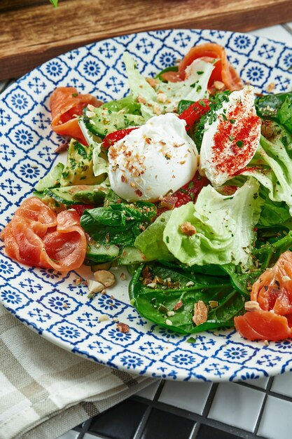 Крупным планом вид на аппетитный салат с лососем, шпинатом, яйцом-пашот и рикоттой в красивой синей керамической пластине на белой поверхности. Вкусная еда. Плоская планировка