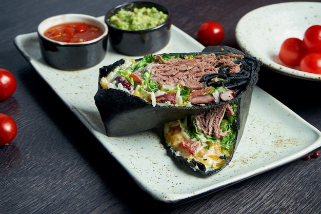 Close up view on burrito with beef, rice, tomatoes, corn and\
bell pepper in black pita on a brown plate with tomato salsa and\
guacamole. vegetarian shawarma roll