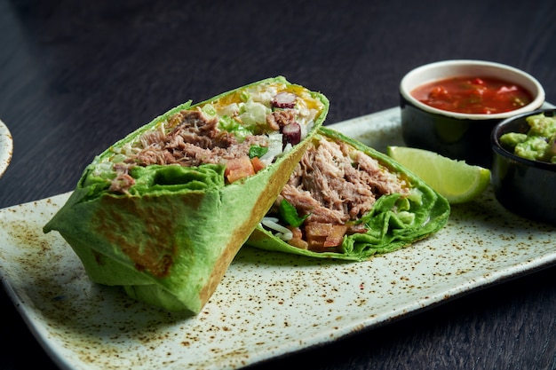 Close up view on burrito with beef, rice, tomatoes, corn and bell pepper in green pita on a brown plate with tomato salsa and guacamole. Vegetarian Shawarma Roll