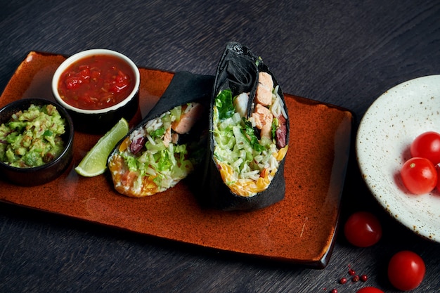 Photo close up view on burrito with salmon, lettuce, rice, tomatoes, corn and bell pepper in black pita on a brown plate with tomato salsa and guacamole. vegetarian shawarma roll