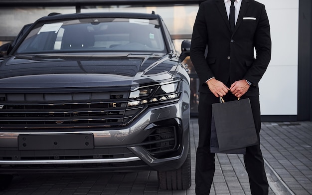 Photo close up view of businessman in black suit and tie stands near modern automobile with shopping bags in hands.